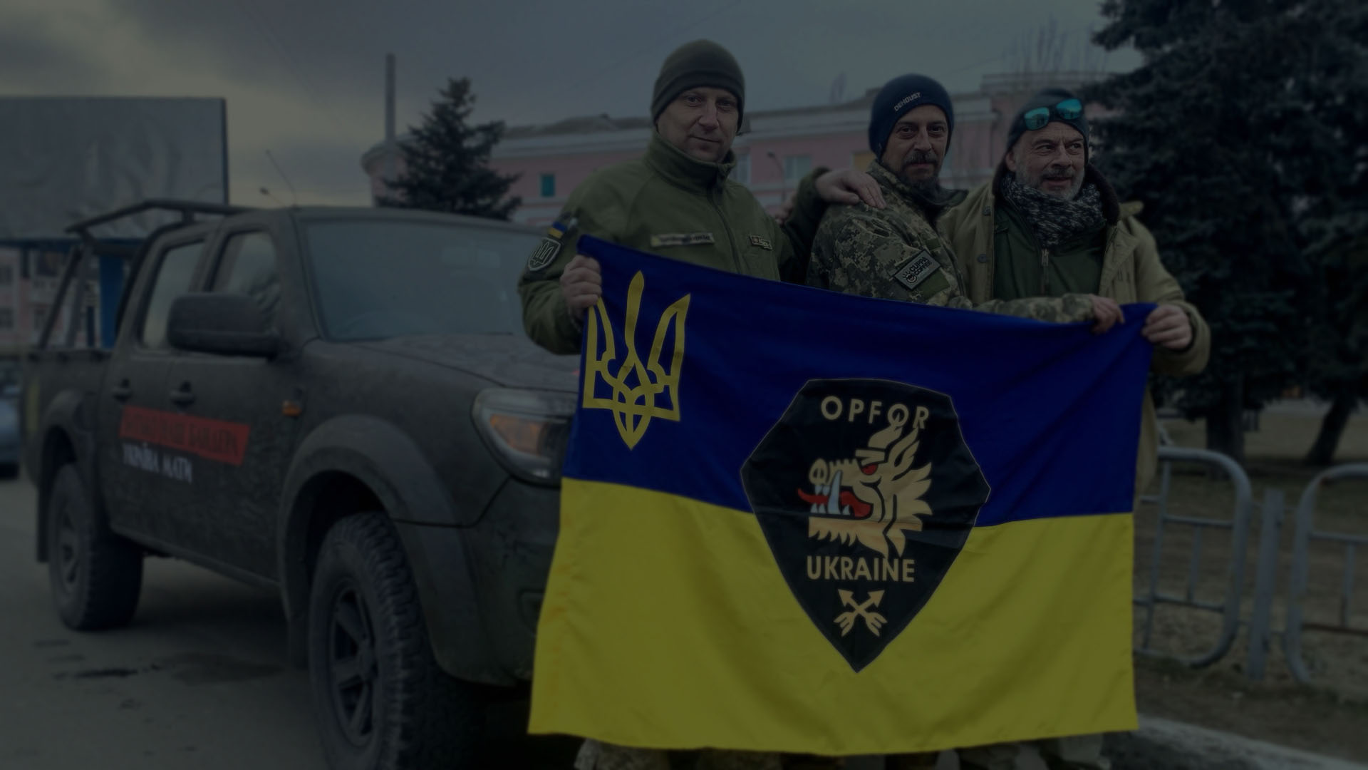 Kaksi UA-sotilasta OPFOR:sta ja Adam pitävät ukrainalaista lippua sotilaallisen poiminta-auton edessä.