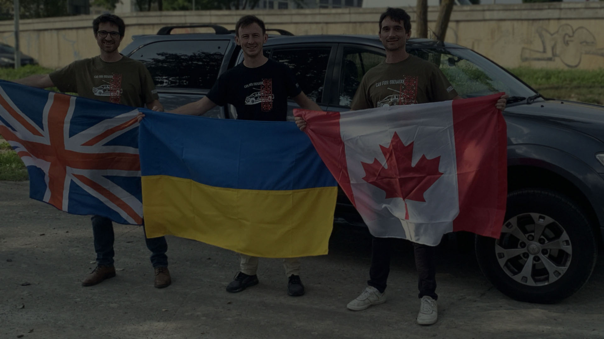 Two brothers and Ivan Oleksii stand in front of two donated SUV pickup trucks holding British, Ukrainian, and Canadian flags.